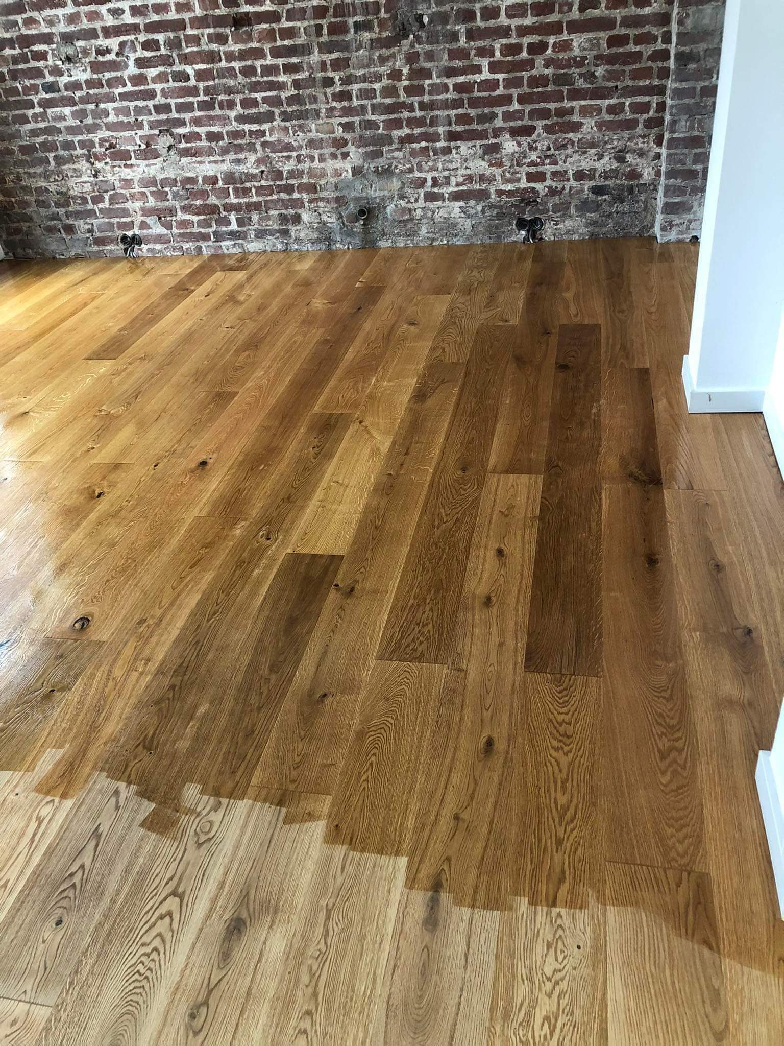 Częściowo odnowiona drewniana podłoga z widoczną różnicą w kolorze i wykończeniu, ceglana ściana w tle.