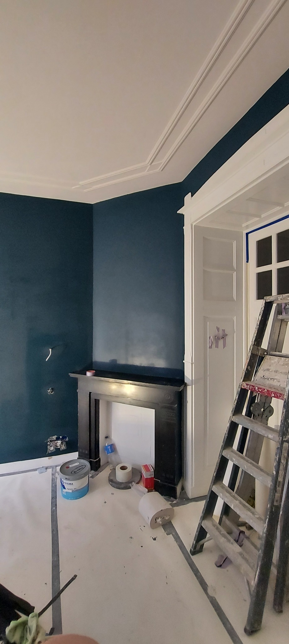 Pokój z pomalowanymi na ciemny granatowo ścianami, z czarnym kominkiem i białą stolarką drzwiową. Na podłodze folia ochronna, widoczna puszka farby Sigma i drabina malarska.
