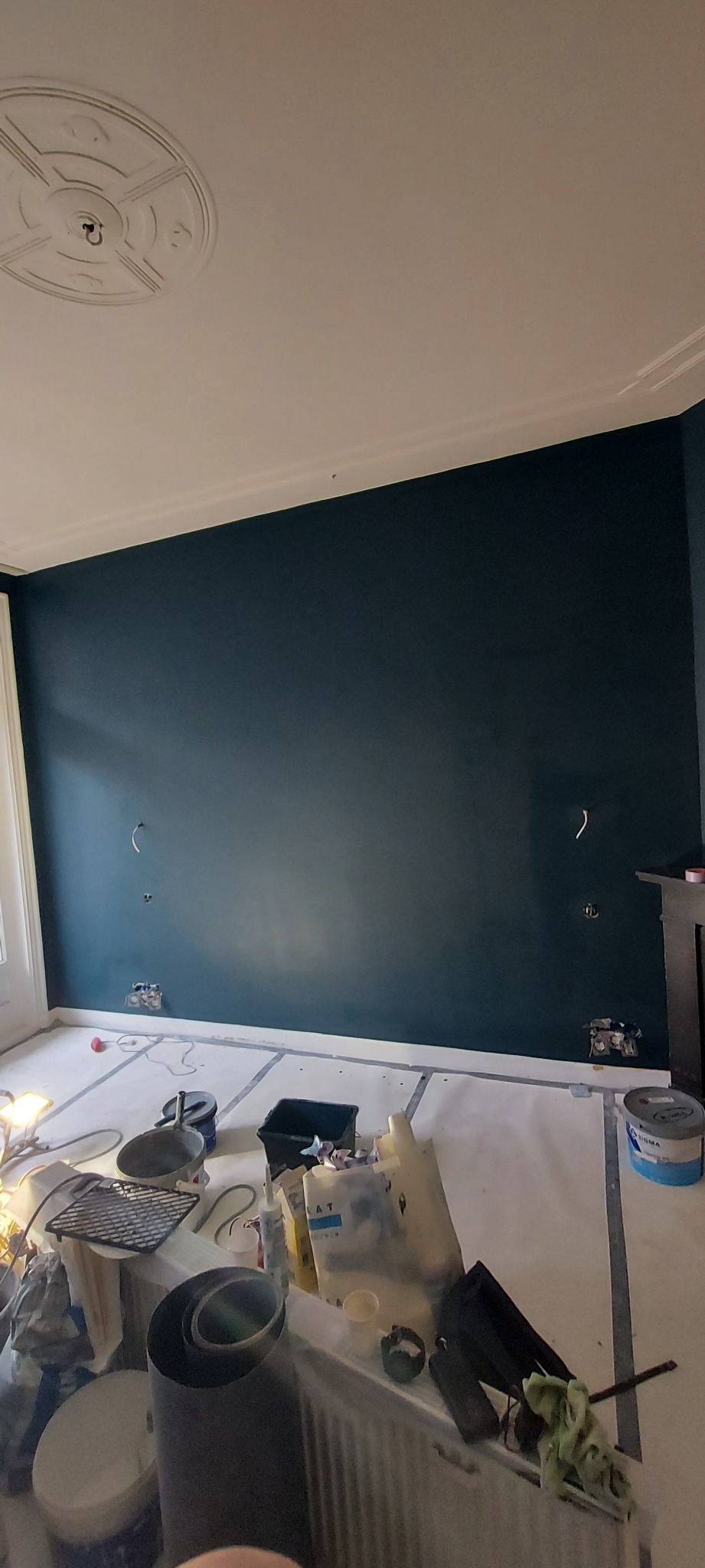 Świeżo pomalowana ściana w odcieniach granatu, zabezpieczona podłoga, widoczne puszki z farbą i narzędzia malarskie, przygotowania do dalszych prac wykończeniowych.