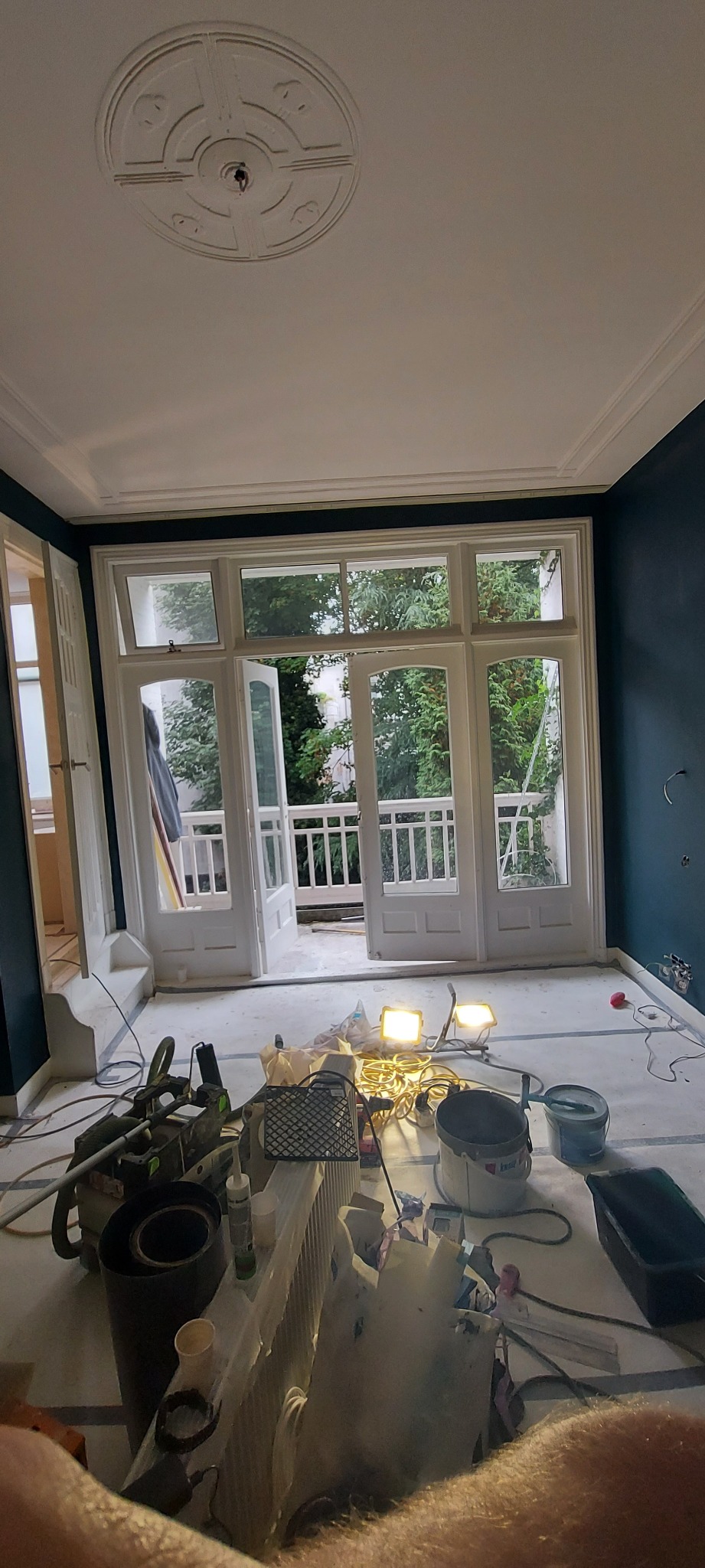 Pokój w trakcie remontu: świeżo pomalowane ciemnoniebieskie ściany, zabezpieczona folią podłoga, otwarte białe drzwi balkonowe z widokiem na zieleń, widoczne narzędzia i akcesoria malarskie, w tym...