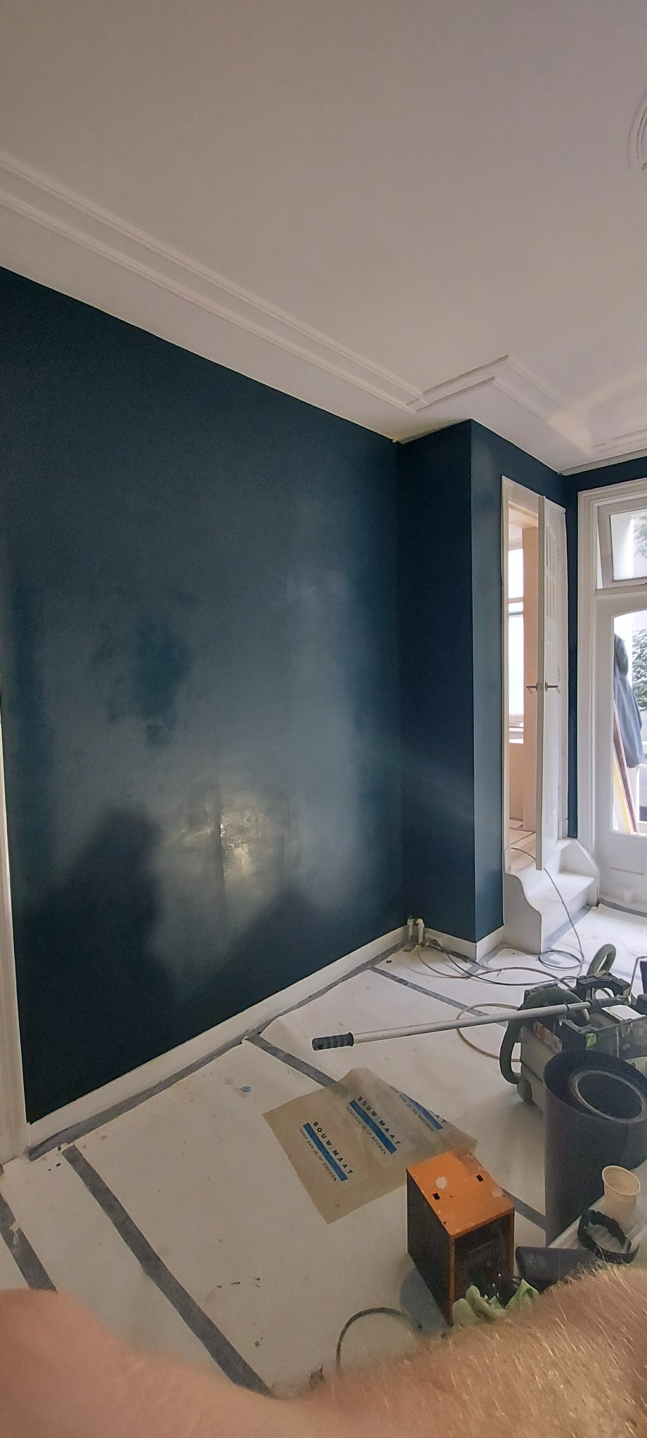 Wnętrze pokoju z pomalowaną na ciemny granatowo-zielony kolor ścianą, widoczne narzędzia malarskie, zabezpieczenia podłogi i listwy sufitowe.
