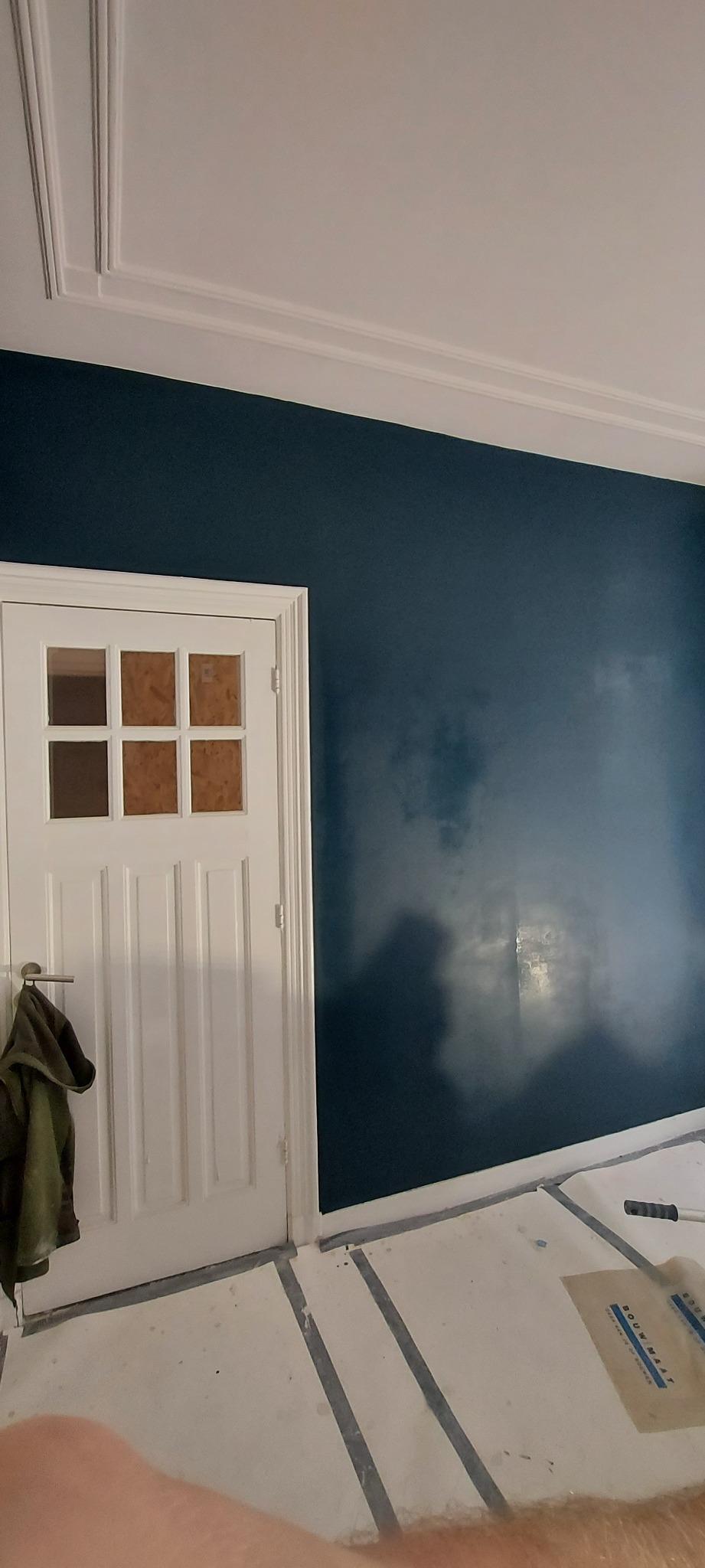 Ściana pomalowana na granatowo z efektem chmur, białe listwy przysufitowe, białe drzwi z szybkami zabezpieczonymi kartonem i kurtka na klamce, podłoga zabezpieczona folią malarską.