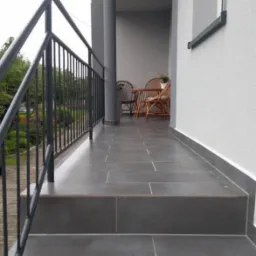 Zadaszony taras z szarymi płytkami, metalową balustradą i rattanowymi meblami w tle.