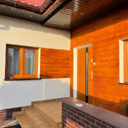 Wejście do domu z elewacją częściowo wykończoną panelami drewnopodobnymi, brązowe okna i drzwi, schody wejściowe z płytek, czerwona dachówka i deska na niskim murku z popielniczką.