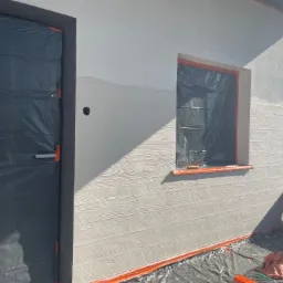 Ocieplanie elewacji domu z imitacją drewna, okno i drzwi zabezpieczone folią, widoczne wiadro z klejem akrylowym Greinplast.