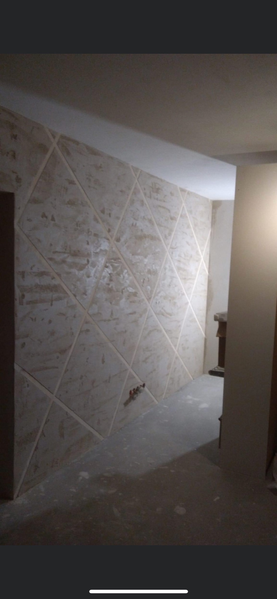 Ściana z dekoracyjną fakturą i geometrycznym podziałem na romby, widoczne rury instalacji wodnej, surowy stan wykończenia podłogi.