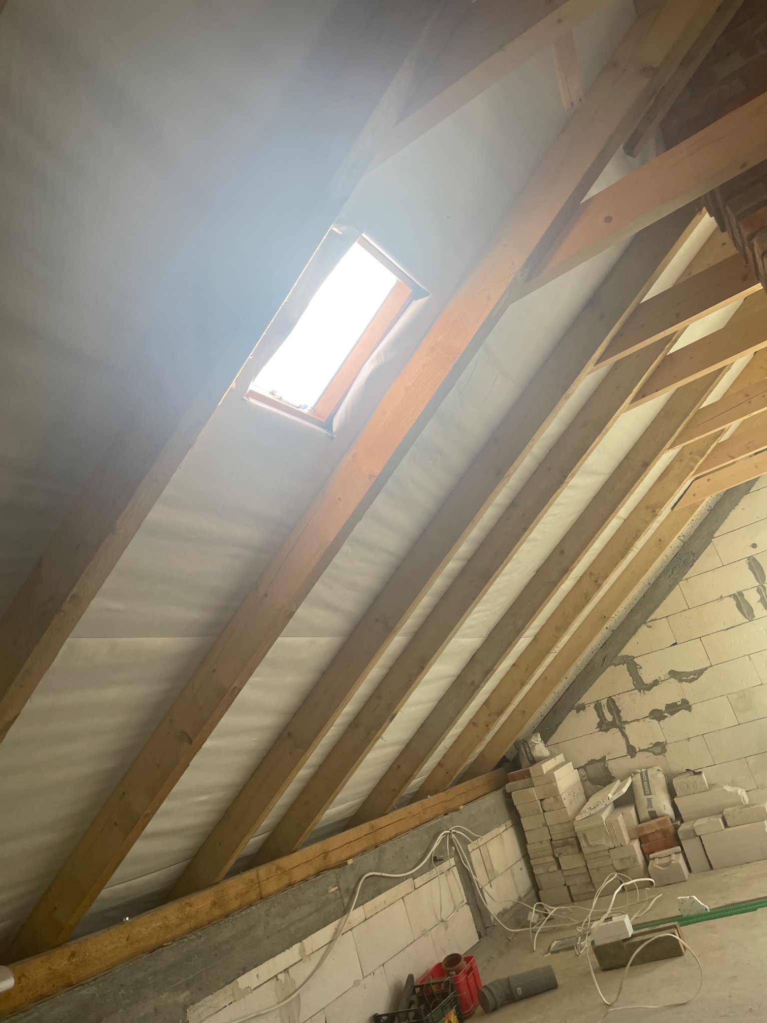 Poddasze w trakcie prac ociepleniowych: widoczne drewniane krokwie, membrana paroizolacyjna, okno dachowe i fragment ściany z bloczków betonowych z przewodami elektrycznymi.
