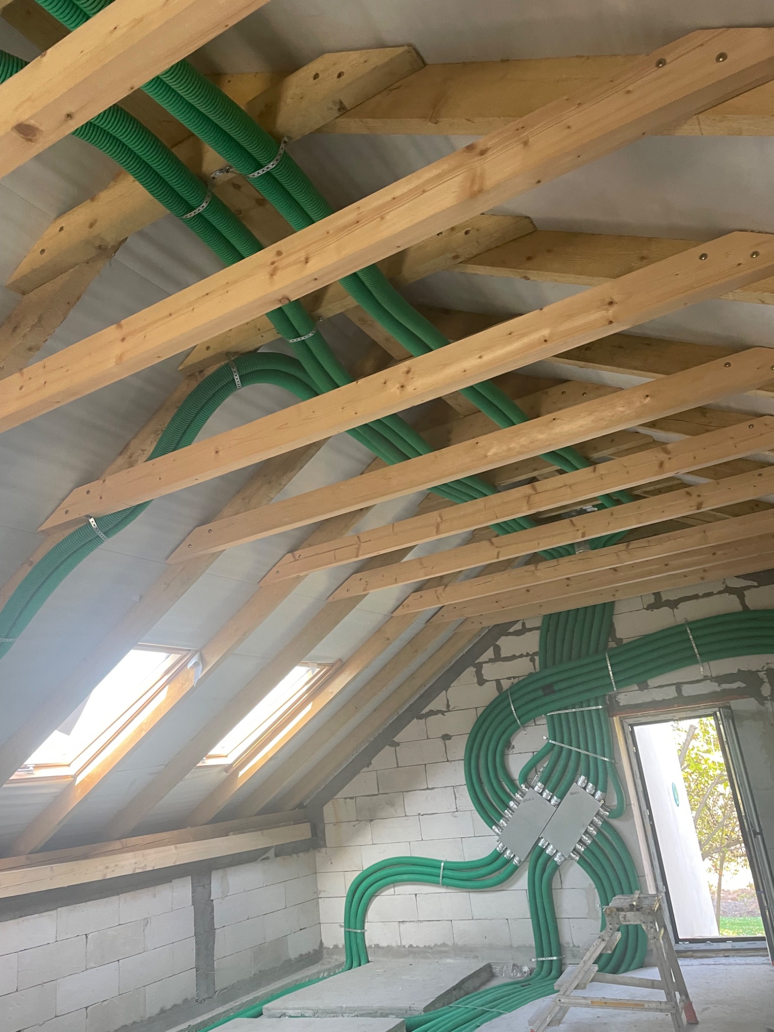 Poddasze w trakcie adaptacji: widoczne krokwie, okna dachowe, ściany z betonu komórkowego oraz liczne zielone rury instalacyjne przymocowane do konstrukcji dachu i ścian.