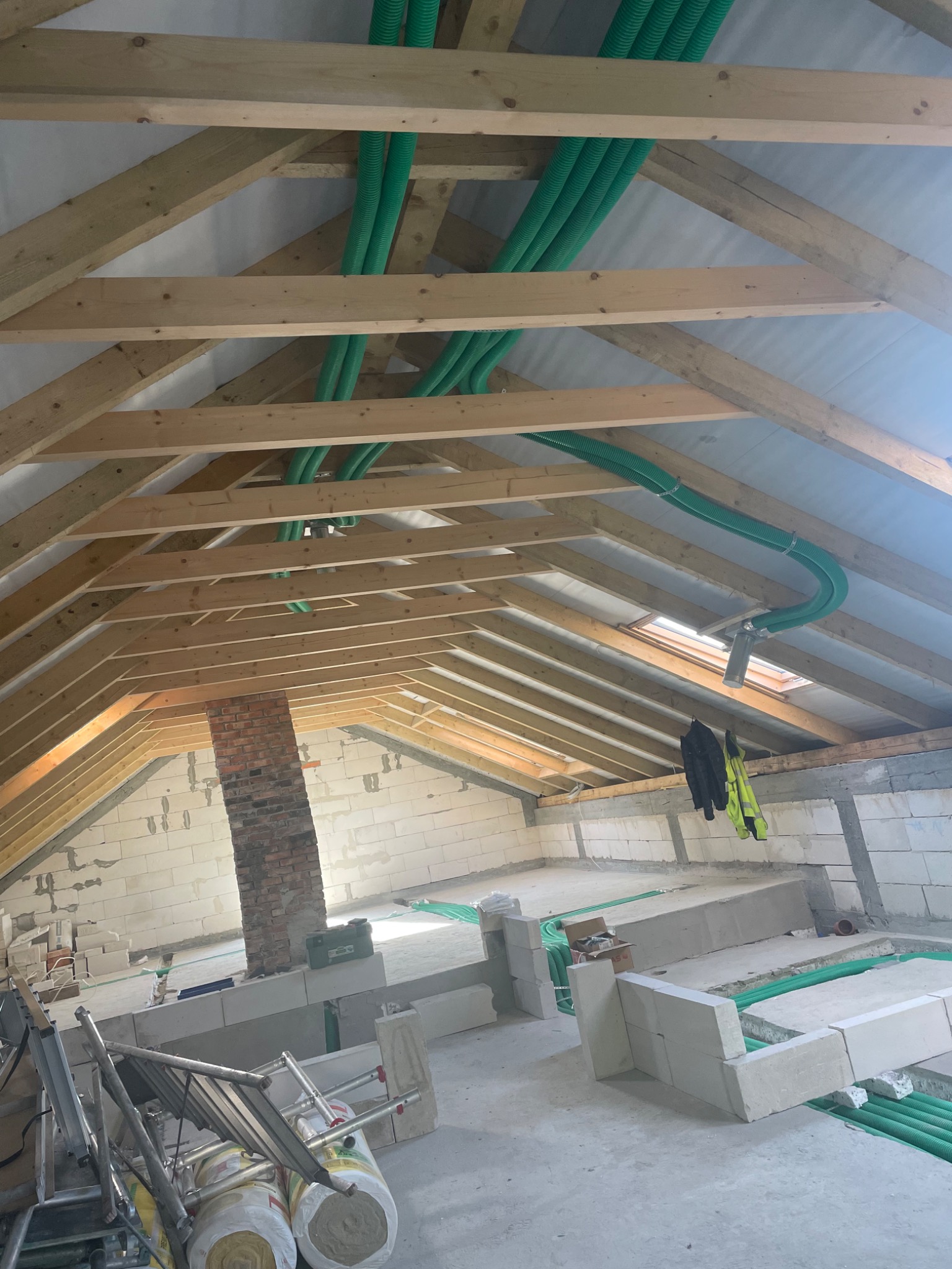 Poddasze w trakcie adaptacji: widoczne drewniane krokwie, izolacja, ceglany komin, instalacja wentylacyjna z zielonych rur oraz bloczki betonowe. Na pierwszym planie złożona drabina i rolki wełny...