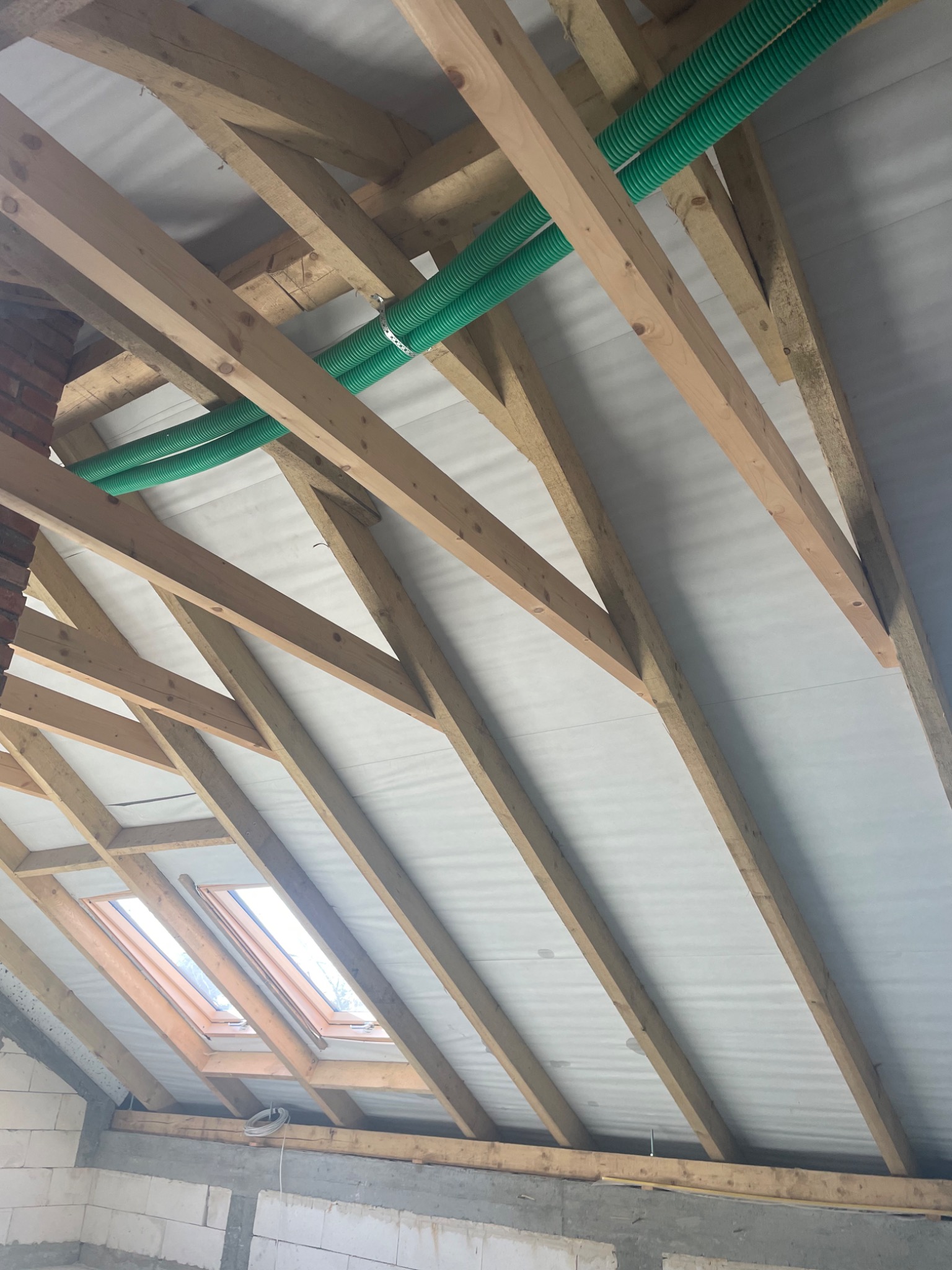 Konstrukcja drewniana poddasza z widocznymi krokwiami, membraną dachową i zielonymi rurami wentylacyjnymi, okno dachowe w tle.