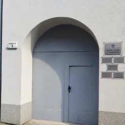 Wejście do budynku z łukowym portalem i szarą bramą, na ścianie tabliczki informacyjne z nazwiskami lekarzy i symbolem medycznym, adres 'Barsary 9' na tabliczce.