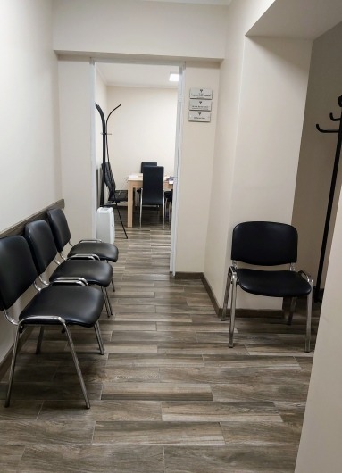 Poczekalnia z czterema czarnymi krzesłami z chromowanymi nogami, podłoga z płytek imitujących drewno, w tle widoczne biuro z krzesłami i stołem, na ścianie tabliczki informacyjne.