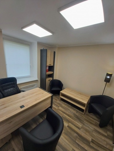 Gabinet psychoterapeuty: drewniany biurko z czarnym krzesłem obrotowym, dwa czarne krzesła dla pacjentów, drewniana szafka RTV, wysoka szafa z półkami, okno z roletą, lampa podłogowa z czarnym...