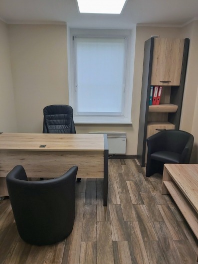 Gabinet z biurkiem o drewnianym wzorze, czarnym krzesłem biurowym i dodatkowym krzesłem dla gości, regał z dokumentami i grzejnik pod oknem.