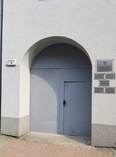 Wejście do budynku z łukowym portalem i szarą bramą, na ścianie tabliczki informacyjne z nazwiskami lekarzy i symbolem medycznym, adres 'Barsary 9' na tabliczce.