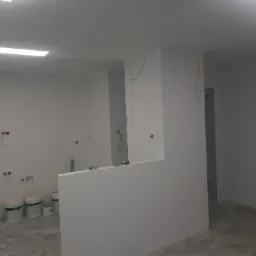 Wnętrze w trakcie prac przygotowawczych w Warszawie: ściany po gładzi, widoczne kable elektryczne wystające ze ścian, białe wiadra z farbą i narzędzia malarskie na podłodze.