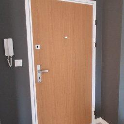 URB GROUP SPÓŁKA Z OGRANICZONĄ ODPOWIEDZIALNOŚCIĄ - Zamknięte, drewniane drzwi wejściowe z wizjerem i srebrną klamką, osadzone w białej futrynie na tle szarej ściany i podłogi z płytek. Widoczny domofon.