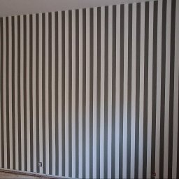 URB GROUP SPÓŁKA Z OGRANICZONĄ ODPOWIEDZIALNOŚCIĄ - Ściana w pionowe pasy w odcieniach szarości i bieli, świeżo pomalowana w mieszkaniu w Poznaniu. Widoczny fragment gniazdka elektrycznego.