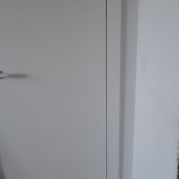 URB GROUP SPÓŁKA Z OGRANICZONĄ ODPOWIEDZIALNOŚCIĄ - Zamknięte, białe drzwi wewnętrzne z widoczną klamką i fragmentem ościeżnicy, na tle białej ściany. Minimalistyczny design, proste linie.