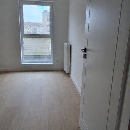 URB GROUP SPÓŁKA Z OGRANICZONĄ ODPOWIEDZIALNOŚCIĄ - Jasne panele podłogowe w pustym pokoju z białymi ścianami, oknem i drzwiami. Widoczny grzejnik. Minimalistyczne wykończenie wnętrza.