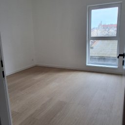 URB GROUP SPÓŁKA Z OGRANICZONĄ ODPOWIEDZIALNOŚCIĄ - Jasne panele podłogowe w pustym pokoju z białymi ścianami i oknem z widokiem na miasto. Minimalistyczne oświetlenie sufitowe.