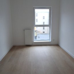 URB GROUP SPÓŁKA Z OGRANICZONĄ ODPOWIEDZIALNOŚCIĄ - Jasne panele podłogowe w pustym, pomalowanym na biało pokoju z oknem i grzejnikiem. Minimalistyczne wnętrze z trzema punktami świetlnymi na suficie.