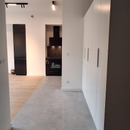 URB GROUP SPÓŁKA Z OGRANICZONĄ ODPOWIEDZIALNOŚCIĄ - Nowoczesne wnętrze mieszkania z szarą podłogą, białą zabudową i czarnymi akcentami oświetleniowymi. Widok na kuchnię z czarnym okapem i sprzętem.