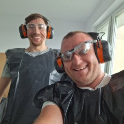 URB GROUP SPÓŁKA Z OGRANICZONĄ ODPOWIEDZIALNOŚCIĄ - Uśmiechnięci pracownicy w okularach ochronnych i słuchawkach, ubrani w czarne peleryny ochronne, gotowi do pracy w jasnym pomieszczeniu.