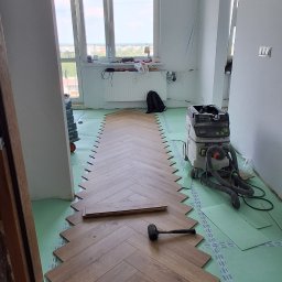 URB GROUP SPÓŁKA Z OGRANICZONĄ ODPOWIEDZIALNOŚCIĄ - Układanie paneli w jodełkę na podkładzie. Widoczny gumowy młotek i odkurzacz przemysłowy. Jasne wnętrze z oknem i widokiem na miasto.