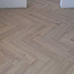 URB GROUP SPÓŁKA Z OGRANICZONĄ ODPOWIEDZIALNOŚCIĄ - Ułożona podłoga z paneli w jodełkę francuską, jasne odcienie drewna, widoczne fragmenty ścian. Nowoczesne wykończenie podłogi w mieszkaniu.