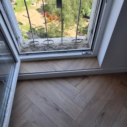 URB GROUP SPÓŁKA Z OGRANICZONĄ ODPOWIEDZIALNOŚCIĄ - Ułożona podłoga z paneli w jodełkę, widok z góry, przy oknie z widokiem na plac zabaw. Jasne panele, biała listwa przypodłogowa.