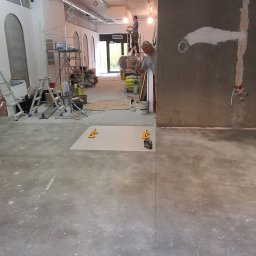 URB GROUP SPÓŁKA Z OGRANICZONĄ ODPOWIEDZIALNOŚCIĄ - Przygotowanie podłoża pod posadzkę w nowym lokalu. Widoczne narzędzia i materiały budowlane, w tle pracownik na drabinie. Surowy stan wykończenia.