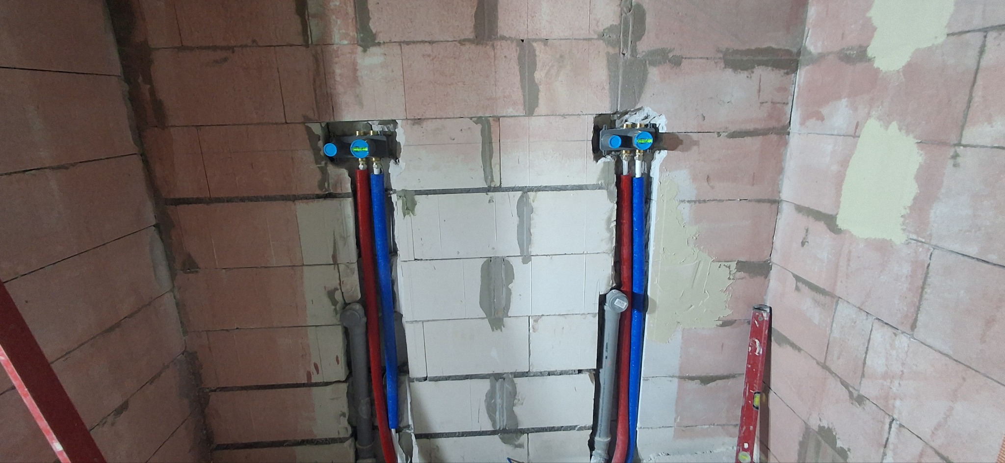 Instalacja wodno-kanalizacyjna w surowej łazience z bloków, z widocznymi rurami w kolorach czerwonym i niebieskim oraz poziomica przy ścianie.