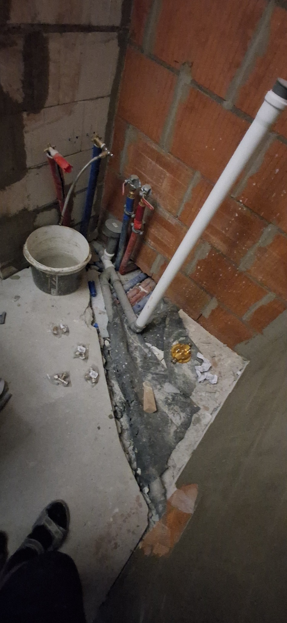 Fragment łazienki w trakcie remontu: widoczne rury instalacji wodnej, wiadro i części armatury na surowej posadzce przy ścianie z cegły. Ujęcie z góry.