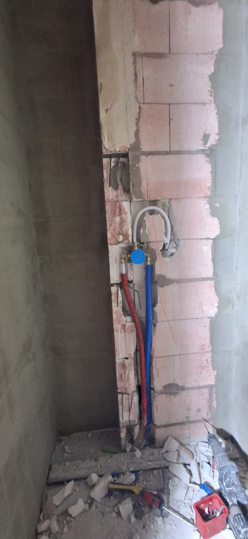 Montaż instalacji wodnej w łazience w trakcie remontu. Widoczne czerwone i niebieskie rury, fragment ściany z cegły oraz narzędzia.