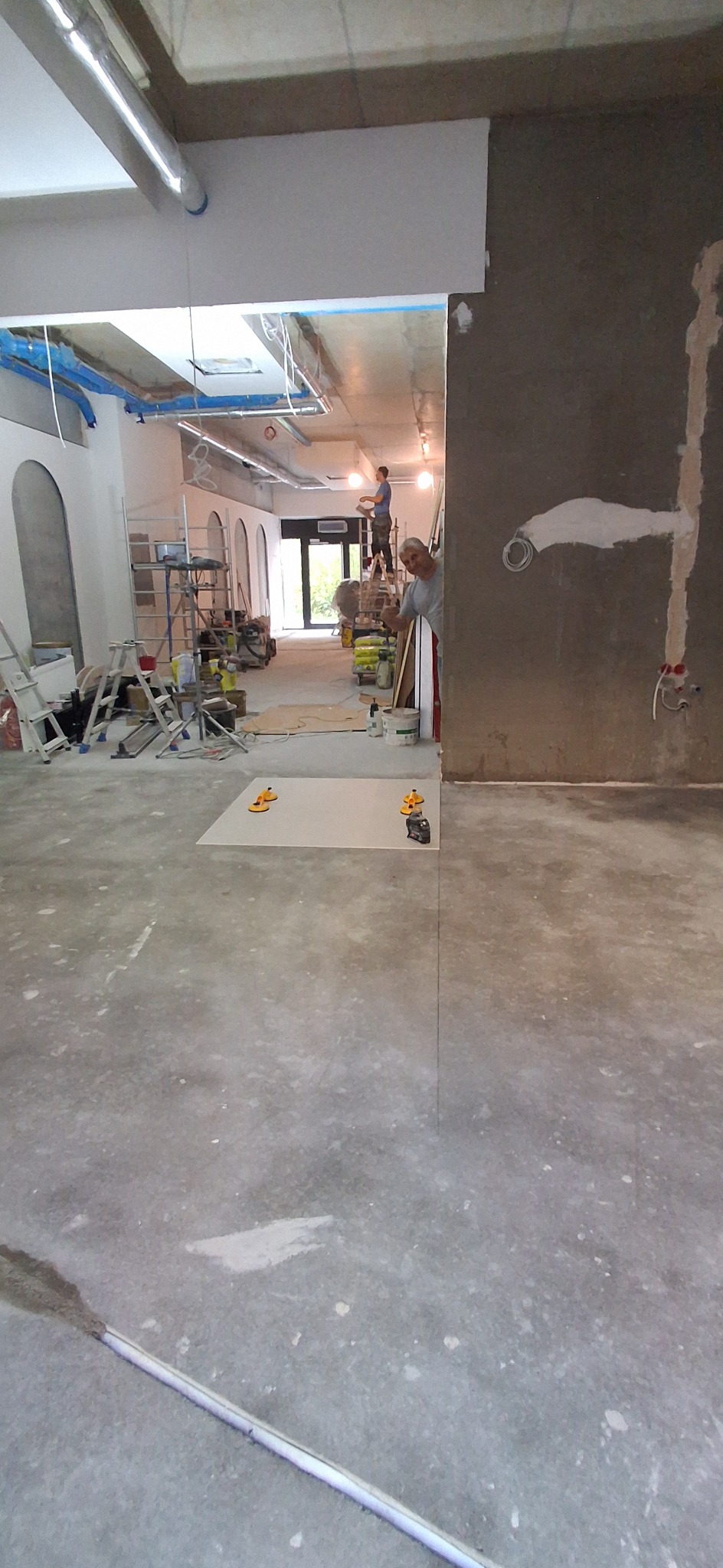 Wnętrze w trakcie prac wykończeniowych: szpachlowanie ściany, układanie płyty z przyssawkami na podłodze, drabiny i narzędzia w tle. Widoczni dwaj pracownicy.