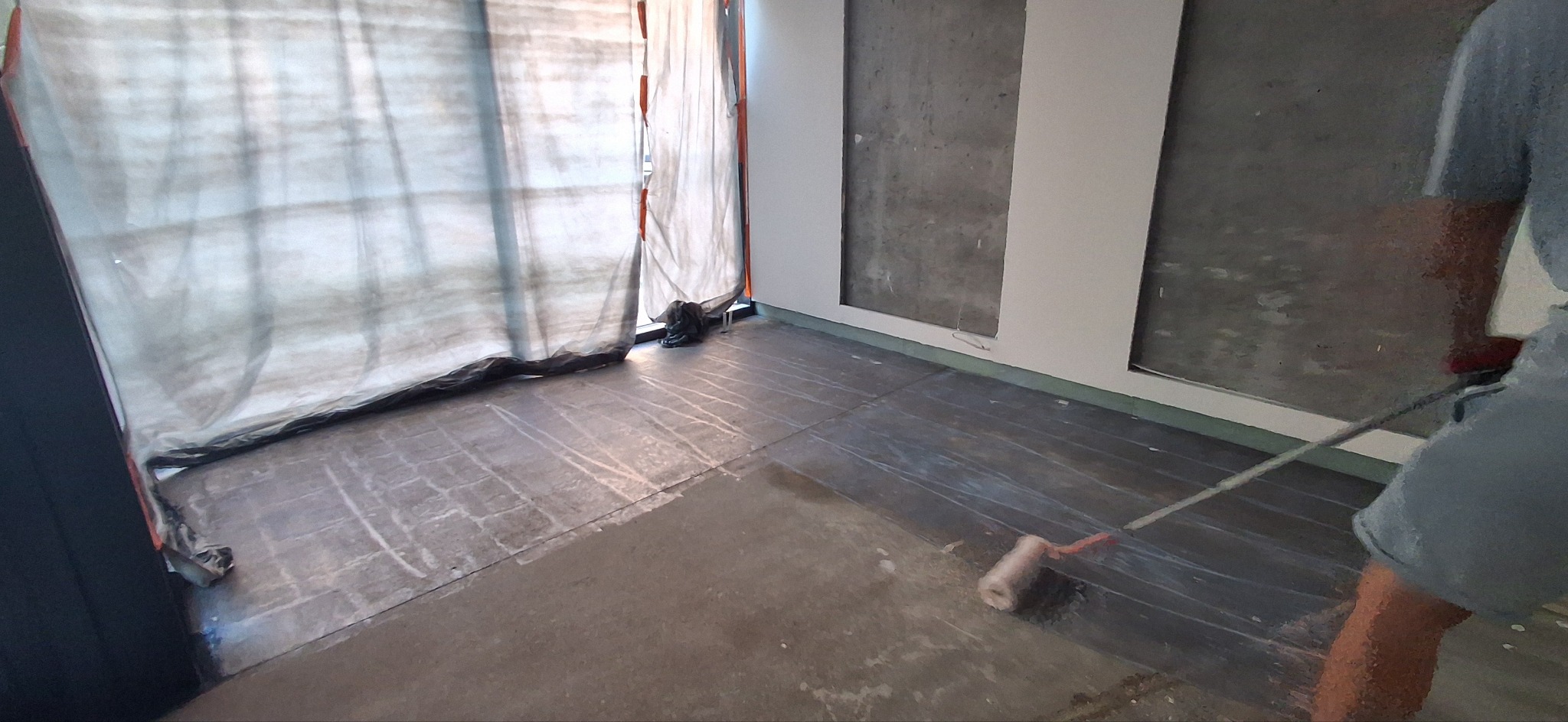 Przygotowanie podłoża: wałkiem nakładana jest warstwa preparatu gruntującego na zabezpieczoną folią podłogę w pomieszczeniu z osłoniętym oknem i surowymi ścianami.