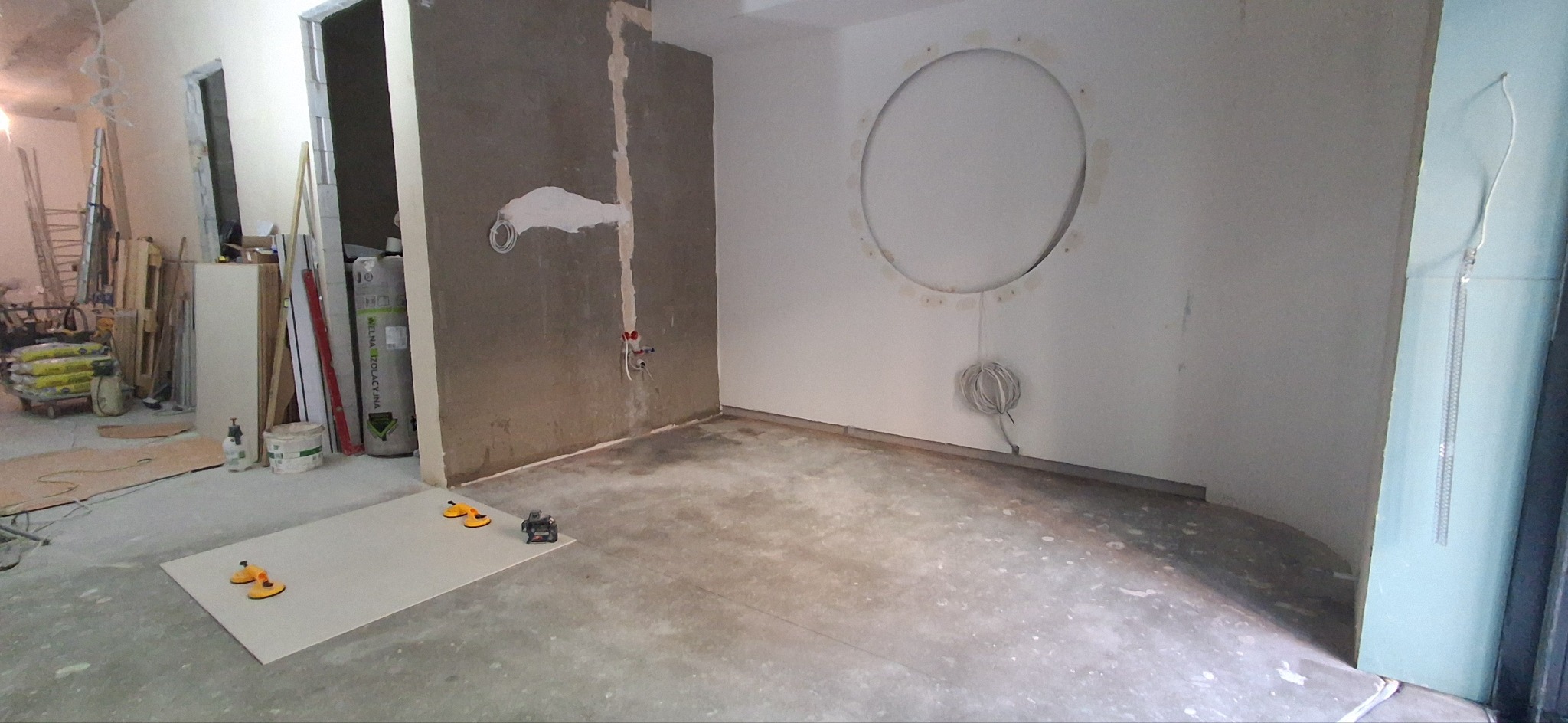 Wnętrze w trakcie prac wykończeniowych: surowa ściana z instalacją, gładka ściana z okrągłym otworem, płyta karton-gips i narzędzia na podłodze. Widoczne materiały budowlane.