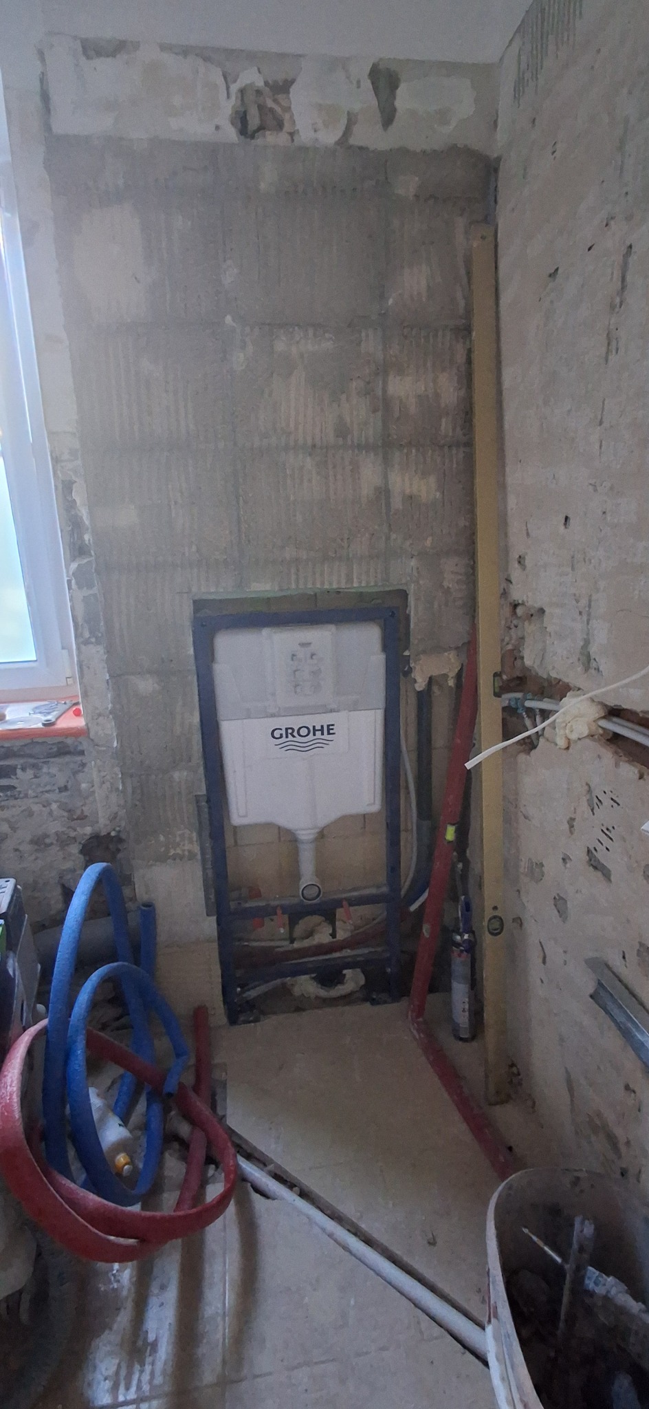 Montaż stelaża podtynkowego Grohe w surowej łazience. Widoczne instalacje hydrauliczne, poziomica i narzędzia. Ściany w stanie deweloperskim, przygotowane do dalszych prac.