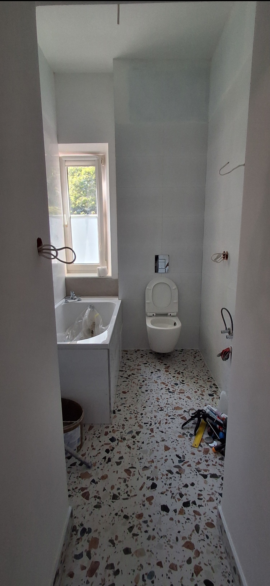 Jasna łazienka w trakcie wykańczania: białe płytki, mozaika na podłodze, wisząca toaleta. Widoczne narzędzia i instalacje przed ostatecznym montażem.