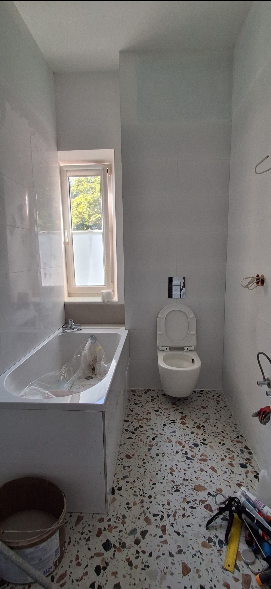 Jasna łazienka po remoncie z mozaikową podłogą, białą armaturą i oknem. Widoczne narzędzia i materiały budowlane sugerują etap wykończeniowy.