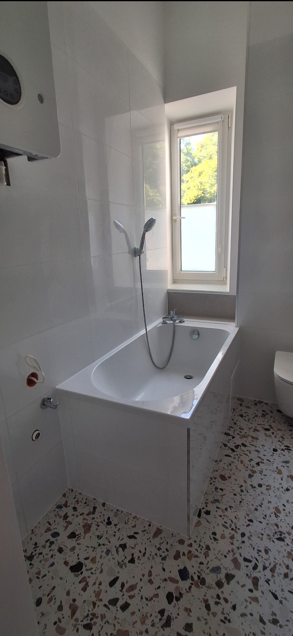 Biała wanna z prysznicem w wyremontowanej łazience z mozaikową podłogą. Widoczne białe płytki na ścianach i okno z widokiem na zieleń.