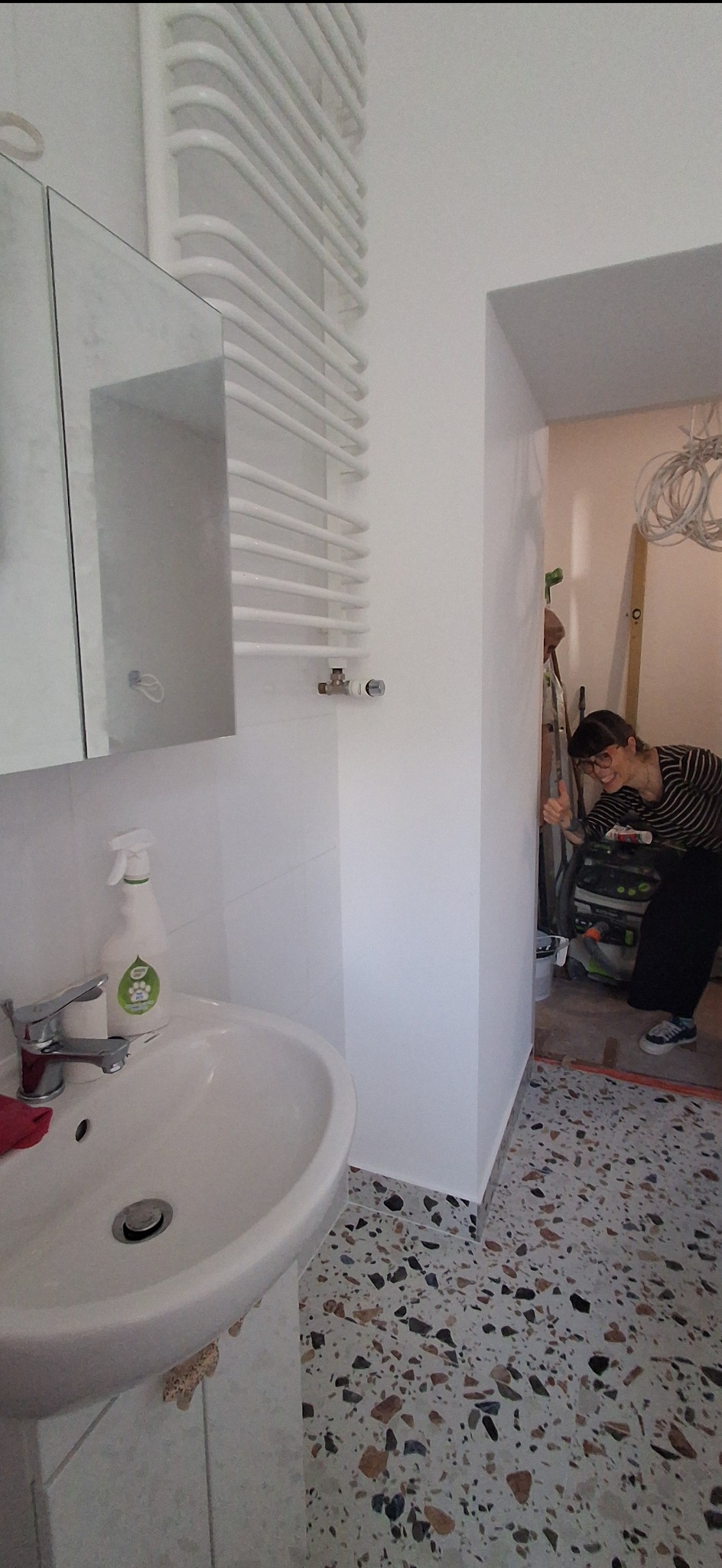 Biała łazienka po odświeżeniu ścian, z umywalką, grzejnikiem drabinkowym i terazzo na podłodze. W tle osoba z podniesionym kciukiem, sugerująca zadowolenie z efektu.