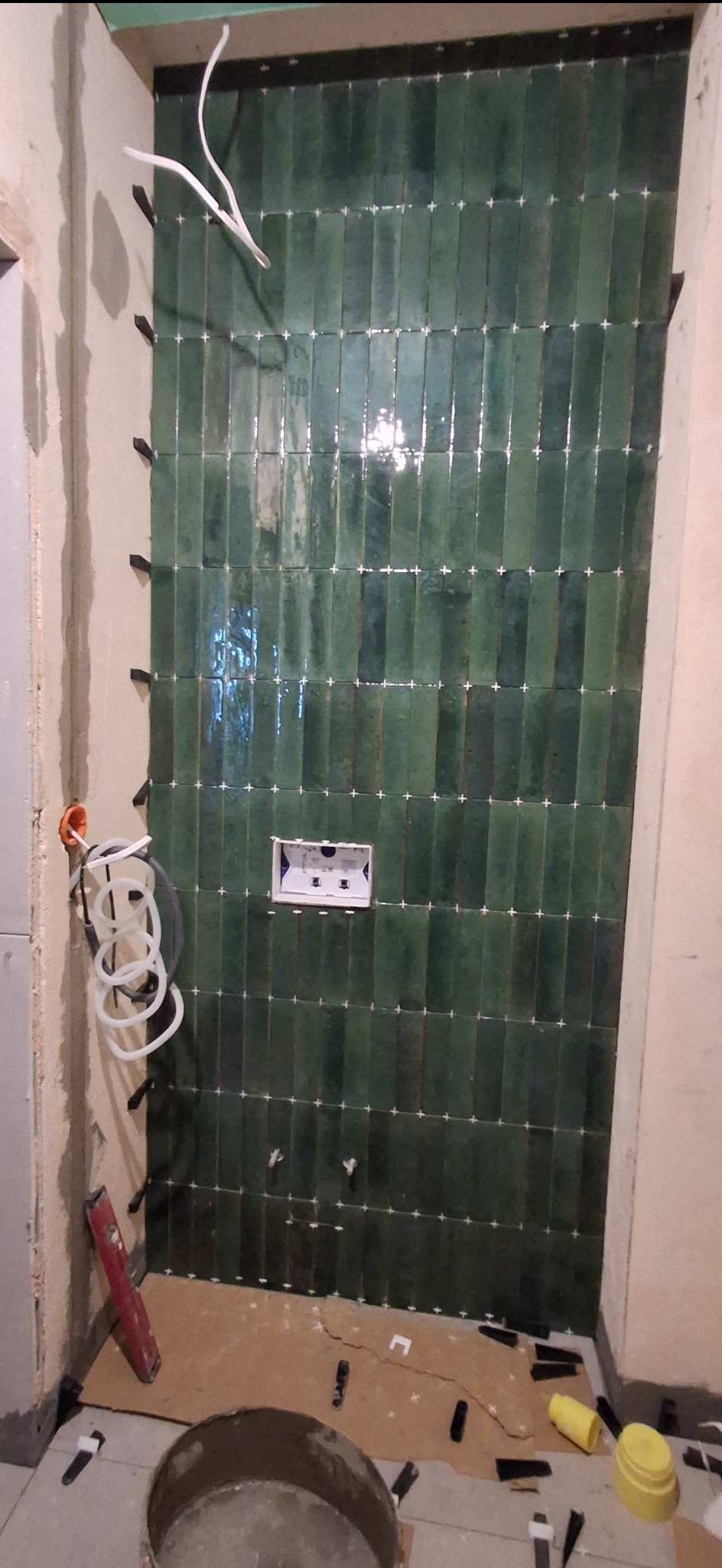 Ściana w łazience z ułożonymi, błyszczącymi, zielonymi płytkami wertykalnymi. Widoczne krzyżyki dystansowe, puszka podtynkową i fragment instalacji.