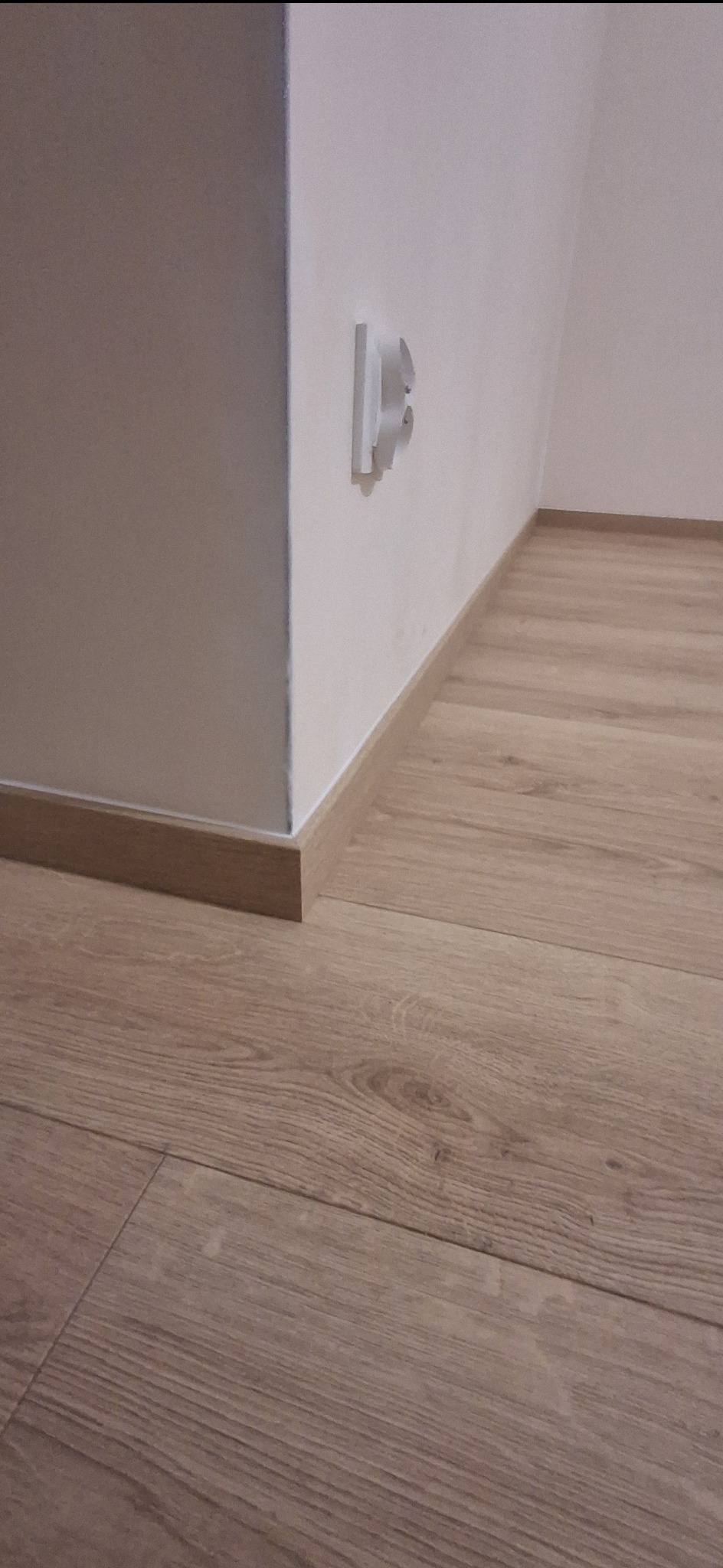 Jasne panele podłogowe z drewnianą listwą przypodłogową przy białej ścianie z gniazdkiem elektrycznym. Ujęcie z niskiej perspektywy.