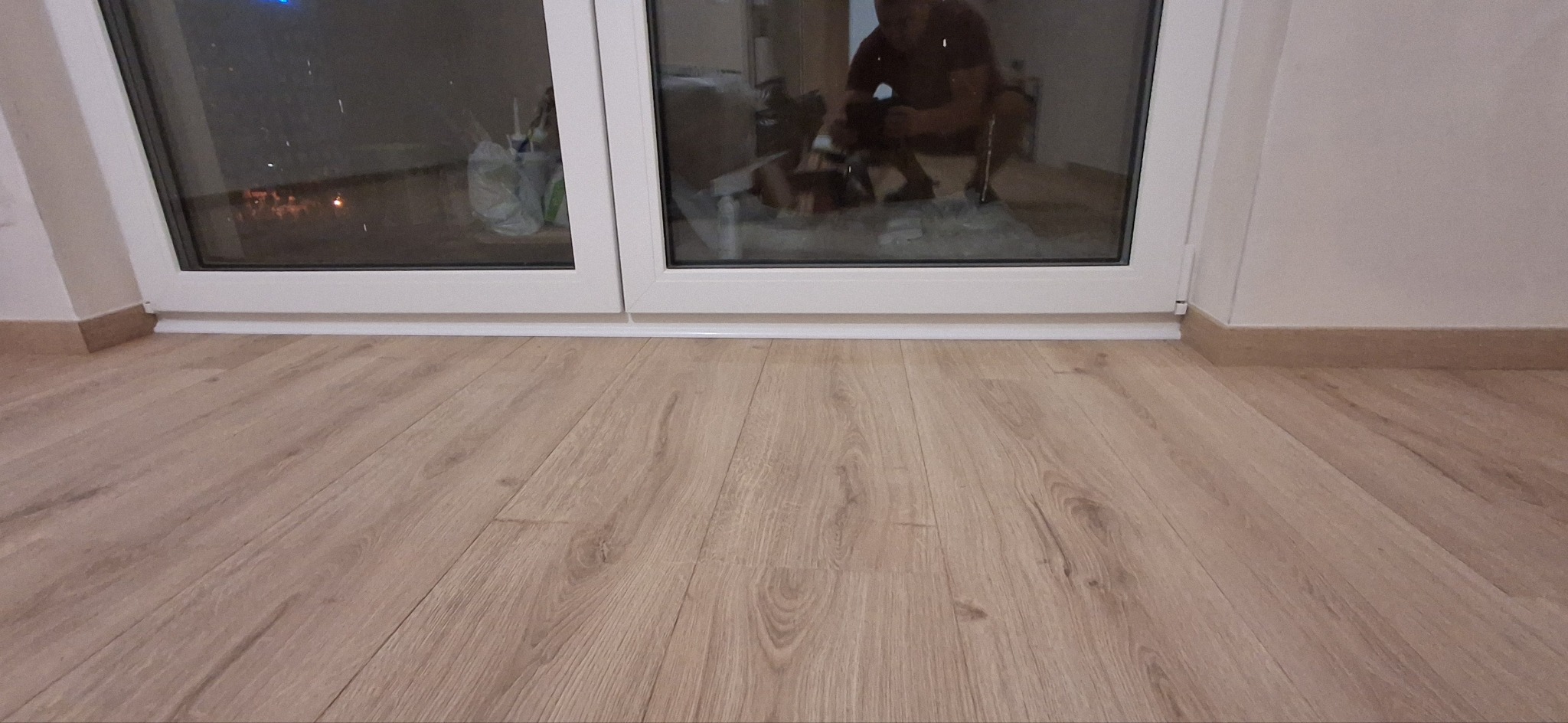 Jasne panele podłogowe ułożone przy białym oknie z widocznym odbiciem osoby wewnątrz, listwy przypodłogowe w kolorze drewna.