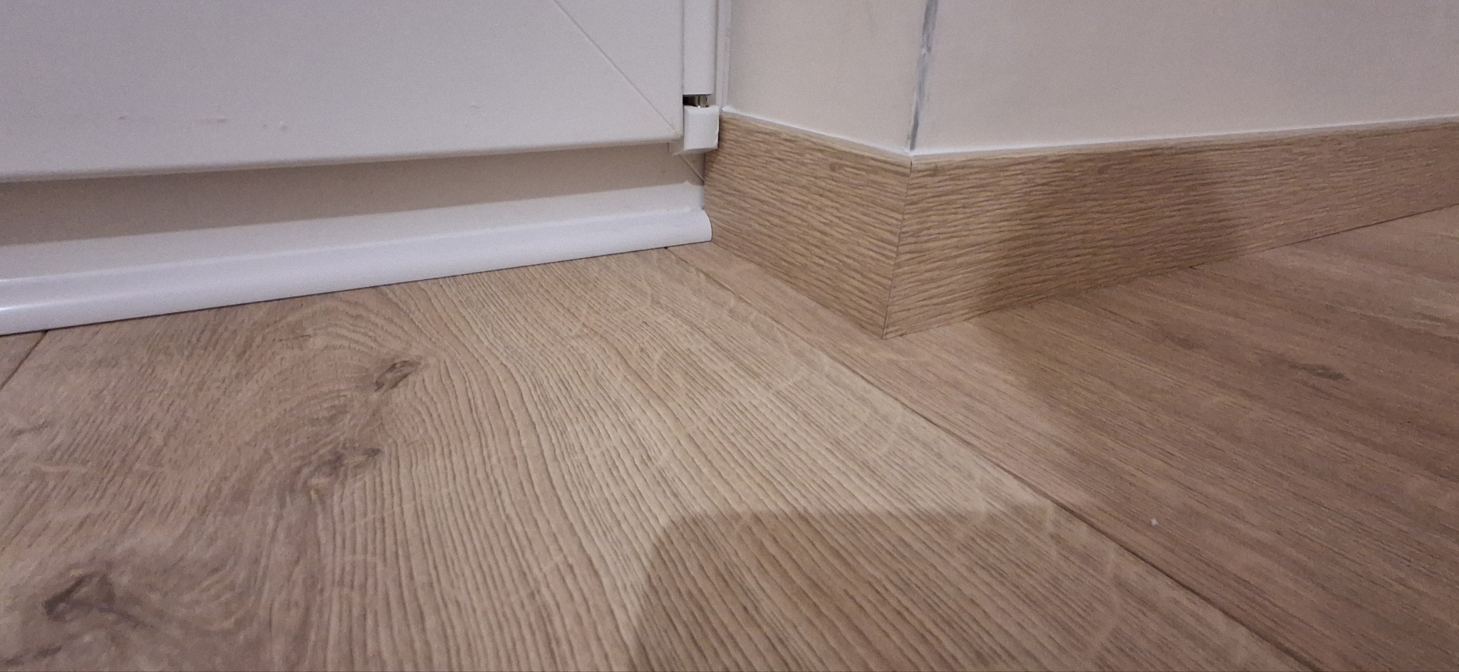 Jasne panele podłogowe z drewnopodobną listwą przypodłogową i fragmentem białej ramy okiennej w rogu pomieszczenia. Detal wykończenia wnętrza.