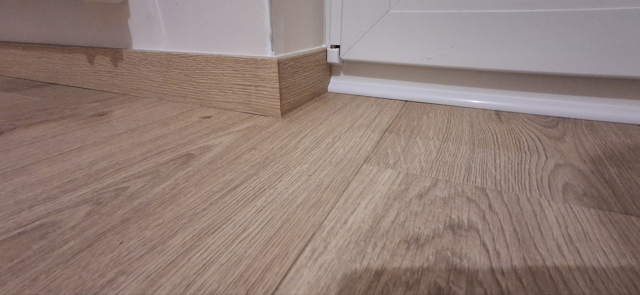 Jasne panele podłogowe z drewnopodobną listwą przypodłogową i białą listwą przy oknie, ukazane z niskiego kąta.