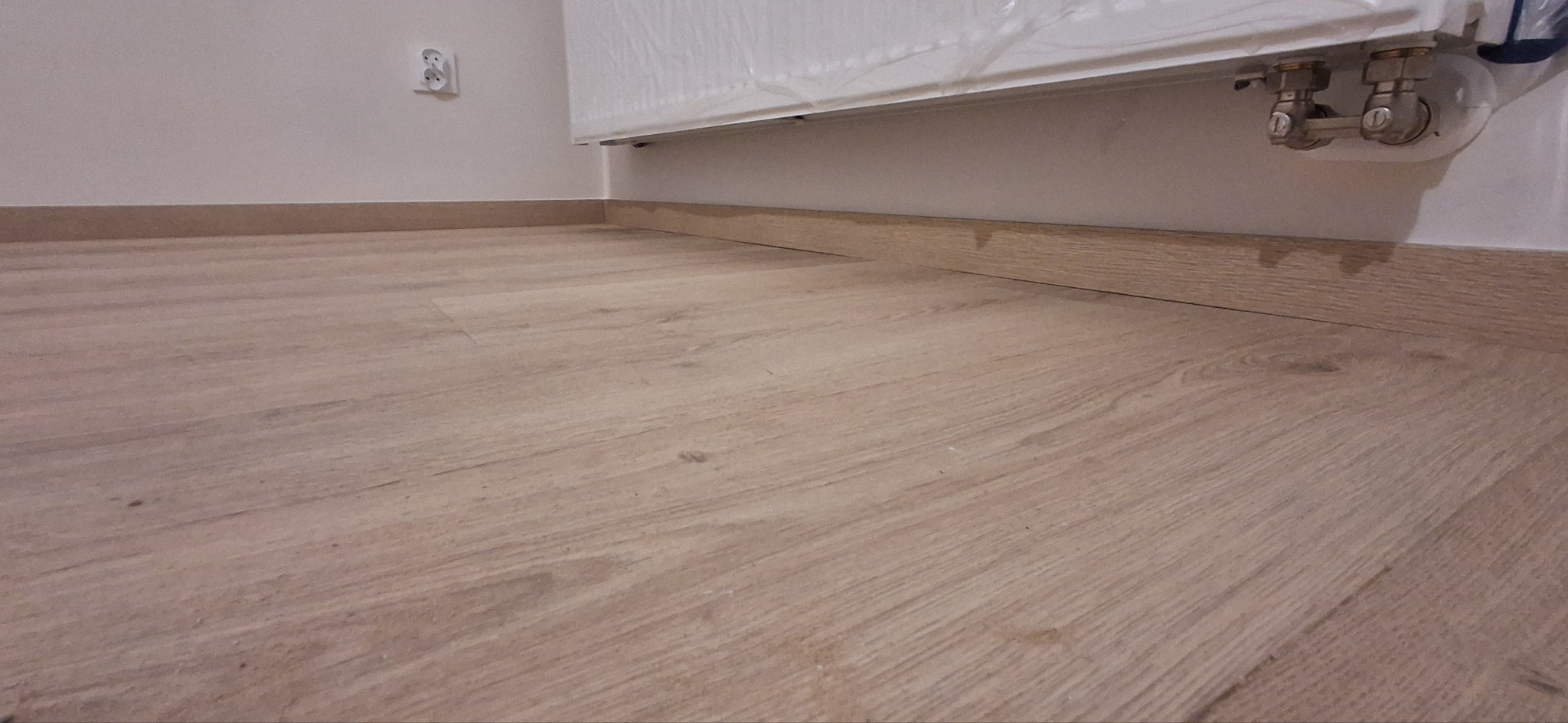 Jasne panele podłogowe z drewnopodobną listwą przypodłogową, fragment grzejnika z zaworami. Widoczny kontakt elektryczny na ścianie.