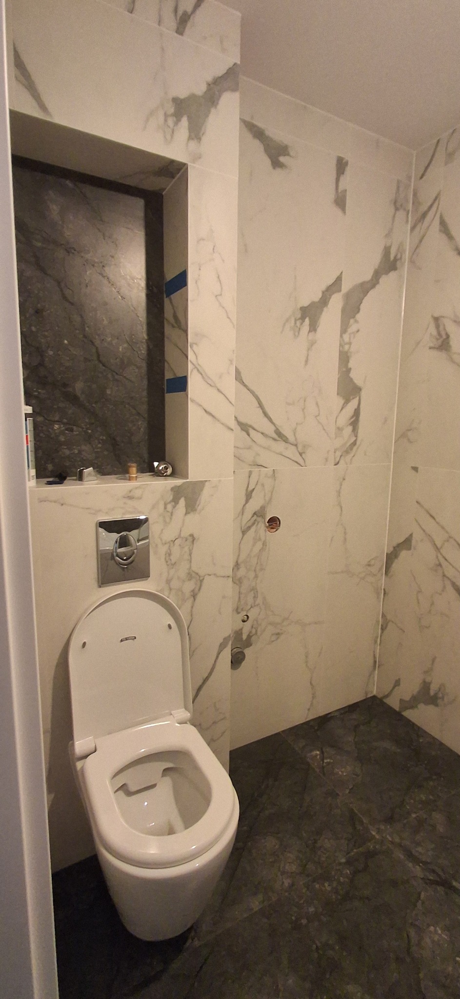 Biała toaleta wisząca w wykończonej łazience z marmurowymi płytkami i wnęką z ciemnego marmuru. Widoczne elementy instalacji.